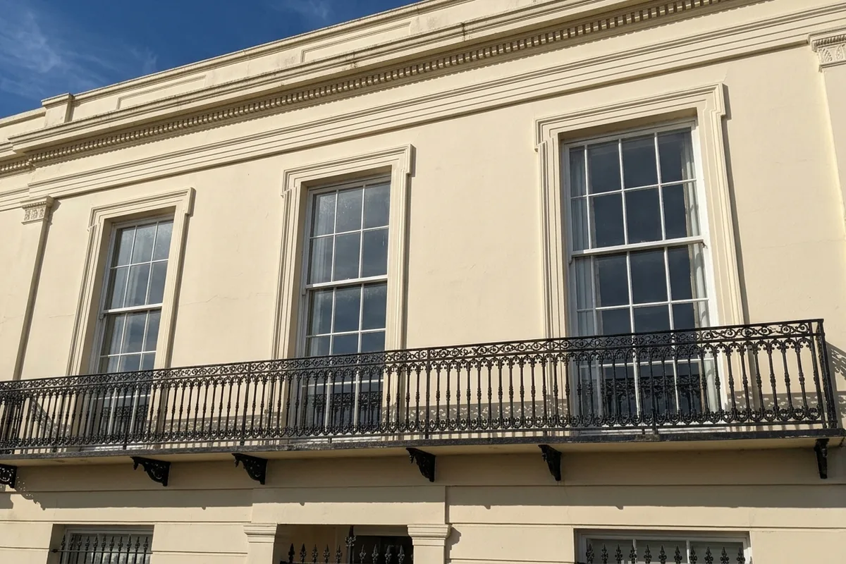 Cream stucco Regency house with tall first-floor sash windows behind a black iron balcony in sunshine